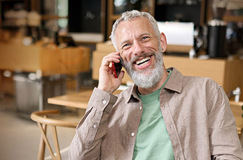 Happy smiling middle aged professional business man, older mature entrepreneur sitting at cafe table talking on cell phone making mobile call remote working online on laptop and having morning coffee.